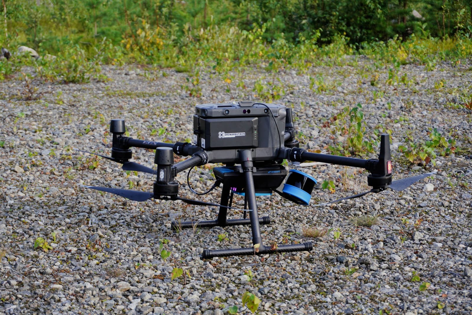 Large quadcopter at rest on gravel with a blue and black LiDAR payload beneath its body