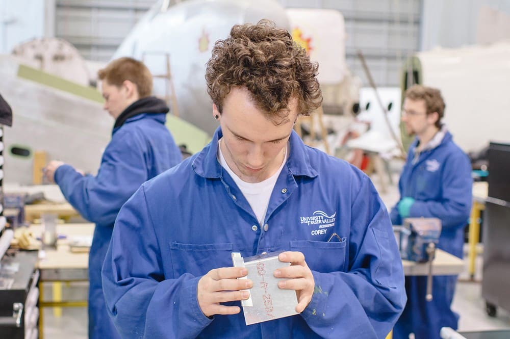 Student in blue workshop coat examining a metal training piece at an aircraft structures program