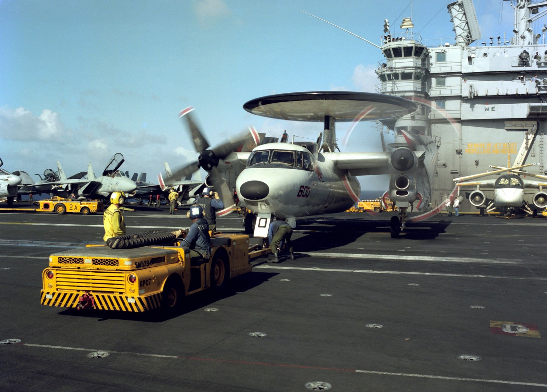 Historical Navy deck crew with a towbar between a tractor and an E2C Hawkeye in 1986